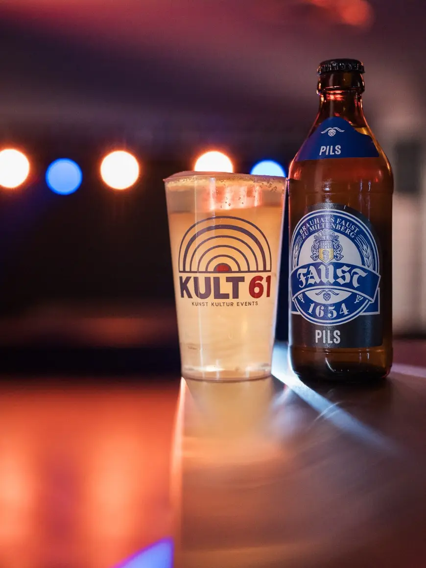 Glas und Flasche Bier mit KULT61-Logo auf der Theke, Bühnenlicht im Hintergrund